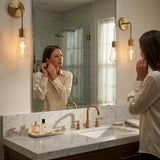 Base up cross filament LED bulbs in brass bathroom vanity sconces beside a mirror
